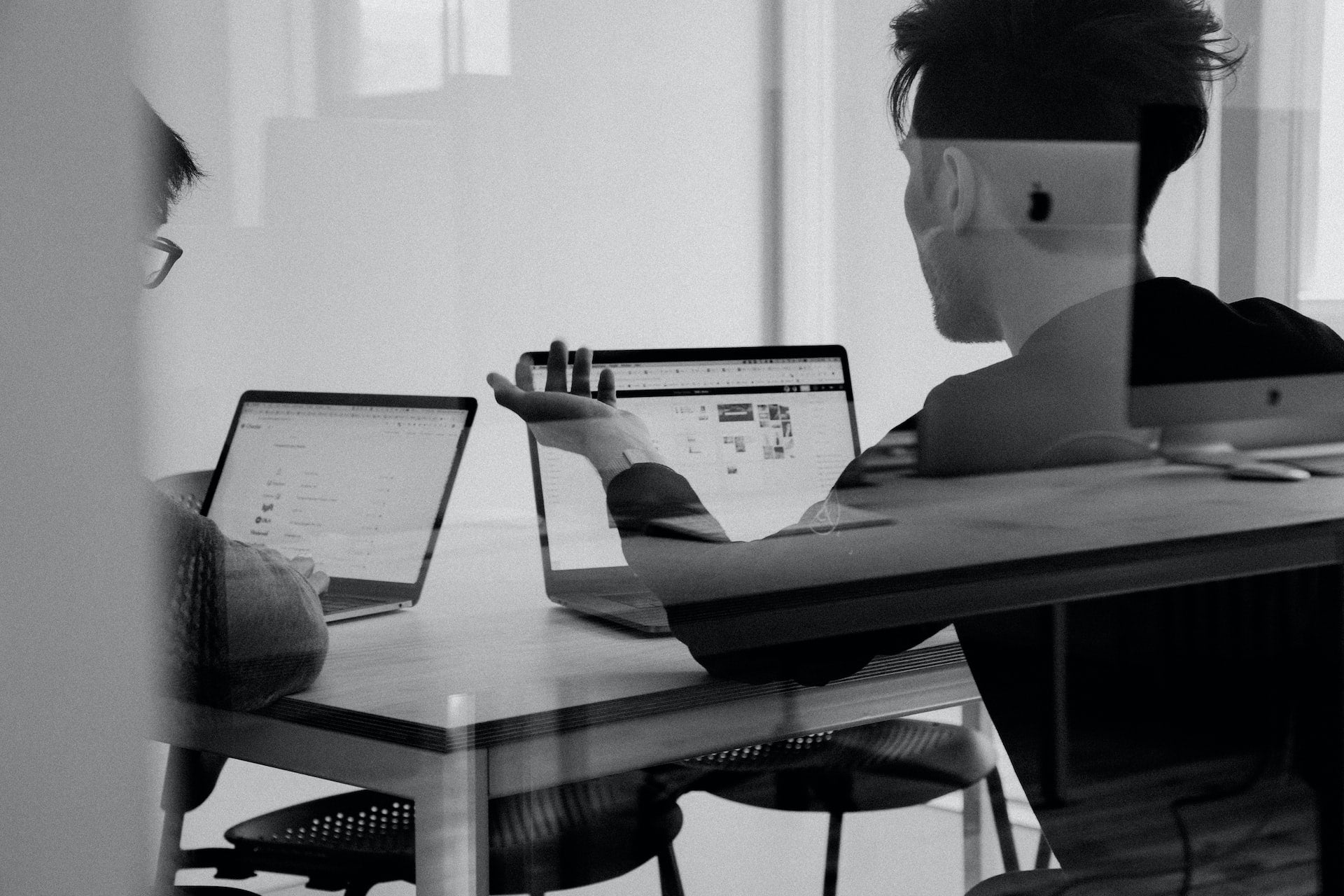 Two people sitting at a table with laptops in a room with large windows.